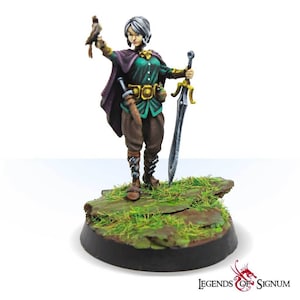 Puede incluir: Una figura miniatura pintada de una guerrera con el pelo corto y plateado. Lleva una túnica verde, pantalones marrones y una capa morada. Sostiene una espada en una mano y un pájaro pequeño en la otra. La figura está de pie sobre una base verde y marrón.
