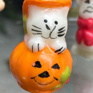 K&ouml;nnte beinhalten: Eine Halloween-Kerze mit einer wei&szlig;en Katze, die auf einem orangefarbenen K&uuml;rbis mit einem geschnitzten K&uuml;rbisgesicht sitzt. Die Katze hat schwarze Schnurrhaare und Augen. Die Kerze hat einen orangenfarbenen Deckel und einen Docht.