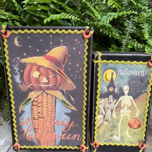 May include: Two rectangular wooden signs with black edges and yellow rickrack trim secured by orange buttons. The larger sign features a corn-headed figure in a hat with the text A Merry Halloween. The smaller sign shows a skeleton in a graveyard.