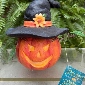 May include: A carved orange pumpkin figurine with a smiling face, wearing a black witch hat adorned with an orange ribbon and a yellow flower. A blue product tag is attached, featuring the text Kathy Hatch Escape from Pumpkin Patch.