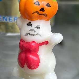 K&ouml;nnte beinhalten: Eine Halloween-Kerze in Form eines Geistes mit einem K&uuml;rbishut und einer roten Schleife. Der K&uuml;rbis ist orange mit einem gr&uuml;nen Stiel und schwarzen Gesichtsz&uuml;gen. Der Geist ist wei&szlig; mit schwarzen Gesichtsz&uuml;gen.