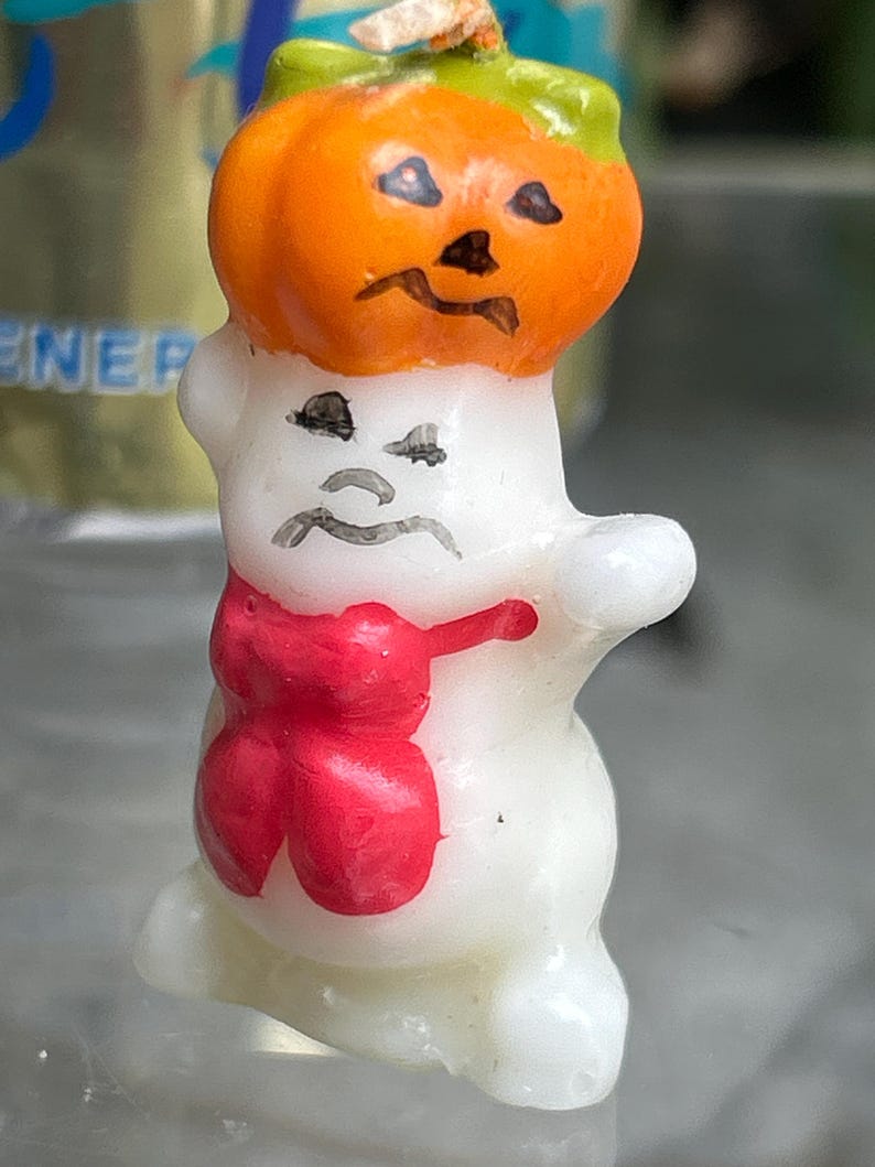 K&ouml;nnte beinhalten: Eine Halloween-Kerze in Form eines Geistes mit einem K&uuml;rbishut und einer roten Schleife. Der K&uuml;rbis ist orange mit einem gr&uuml;nen Stiel und schwarzen Gesichtsz&uuml;gen. Der Geist ist wei&szlig; mit schwarzen Gesichtsz&uuml;gen.