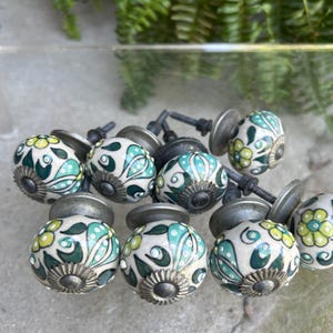 May include: A collection of decorative ceramic cabinet knobs. The knobs are white with a floral design in shades of green, teal, and yellow. Each knob has a silver-toned metal base and screw.