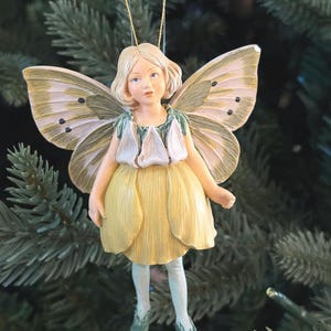 May include: A fairy ornament with a yellow dress and butterfly wings. The fairy has blonde hair, a green top, and light blue shoes. The wings are cream-colored with brown spots. The ornament is hanging from a gold string.