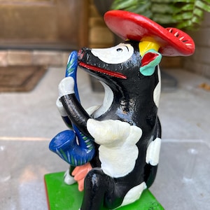 May include: A black and white cow figurine wearing a red hat and playing a blue saxophone. The cow is sitting on a green base with a red trim. The figurine is hand-painted and has a whimsical style.