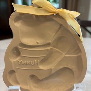 May include: Tan Winnie the Pooh cookie cutter with the word "HONNY" in the center. The cookie cutter has a yellow ribbon tied at the top. The cookie cutter is sitting on a clear plastic stand.
