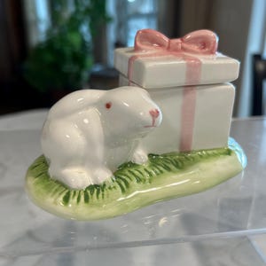 May include: A white ceramic bunny figurine sitting on a green base with a white ceramic gift box with a pink ribbon on top.