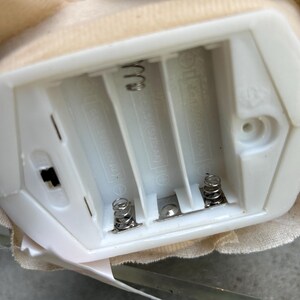 May include: A close-up of a white plastic battery compartment with three slots for AAA batteries. The compartment has metal springs and a small on/off switch. A beige fabric is visible around the compartment.