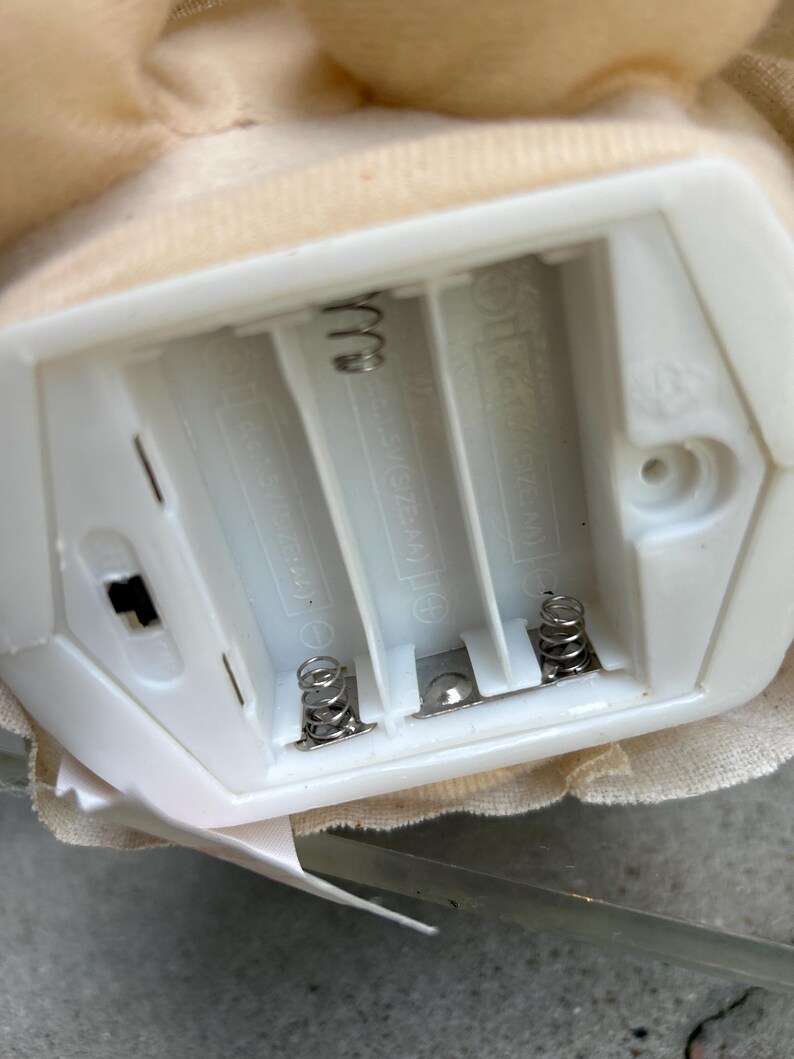 May include: A close-up of a white plastic battery compartment with three slots for AAA batteries. The compartment has metal springs and a small on/off switch. A beige fabric is visible around the compartment.
