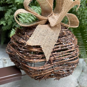 May include: A decorative sphere made of woven brown twigs, adorned with a burlap bow. The sphere has a rustic appearance, with a light dusting of white, and is likely used for home decor or seasonal displays.