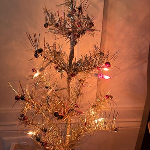 May include: A vintage silver tinsel Christmas tree with small red and black ornaments. The tree is illuminated with small white and pink lights. The base of the tree is a gold-colored, geometric shape.