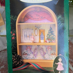May include: A Hallmark Keepsake Ornament box featuring The Nutcracker Ballet display stage. The box shows a two-tiered wooden stage with a ballerina figurine, a Christmas tree, and candy cane decorations. A small ballerina ornament is pictured separately.