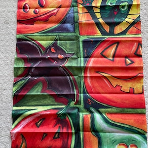 May include: A colorful Halloween-themed fabric panel with a patchwork design. It features a green bat, a black cat, and three orange pumpkins with jack-o'-lantern faces. The background is a mix of blue, green, yellow, and orange.