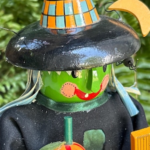 May include: A Halloween witch figurine with a green face, red lips, and a black hat with orange and teal squares. The witch is wearing a black outfit and holding a carved pumpkin. A decorative Halloween item.