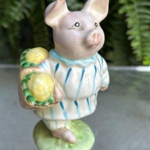 May include: A ceramic figurine of a pig wearing a striped shirt and carrying a basket of yellow lemons. The pig is standing on a green base. The figurine has a light pink face and ears, with blue accents on the shirt.