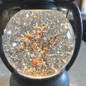 May include: A Halloween-themed snow globe in a black lantern design. Inside, a tree with orange pumpkin faces and a skeleton figure are surrounded by glitter. The globe sits on a black base and has a handle.