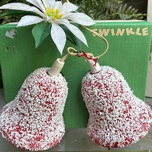 May include: Two red Christmas bell ornaments covered in white glitter. The ornaments are hanging in front of a green box with the word "TWINKLE" in yellow letters. A white poinsettia flower is on top of the box.