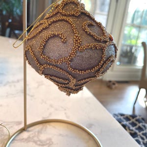 May include: A decorative, diamond-shaped Christmas ornament. The ornament is a muted silver color, embellished with gold-colored beads in a swirling pattern. It hangs from a gold-colored wire and is displayed on a gold-colored stand.