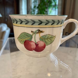 May include: White ceramic teacup with a green and yellow border and a cherry design. The cherries are red with green leaves.