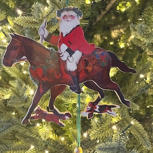 May include: A flat, decorative Christmas ornament featuring a vintage-style illustration of Santa Claus in a red coat riding a brown horse. The horse wears a green wreath with a red bow. A small dog and a fox run below the horse. A green string hangs down.