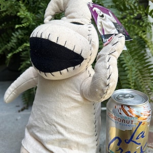 May include: A beige Oogie Boogie plush toy with black stitched details and a large mouth. The doll is holding a tag and is next to a can of La Croix sparkling water. The doll is made of a light beige fabric.