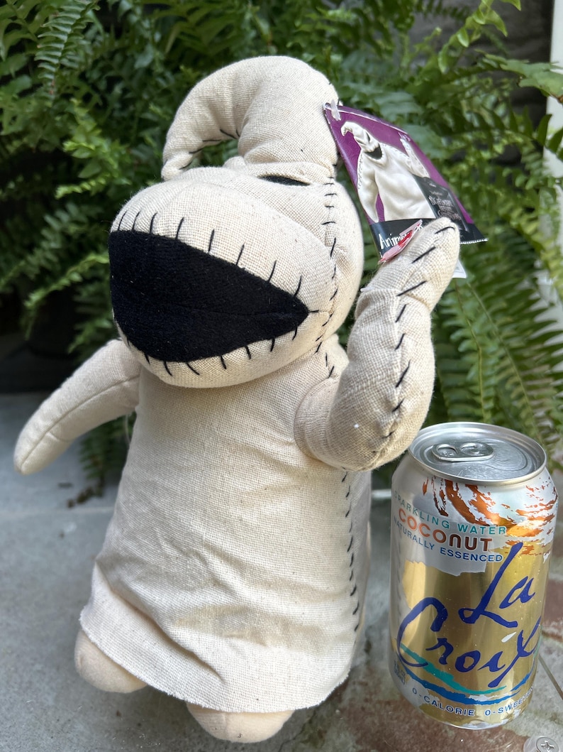 May include: A beige Oogie Boogie plush toy with black stitched details and a large mouth. The doll is holding a tag and is next to a can of La Croix sparkling water. The doll is made of a light beige fabric.