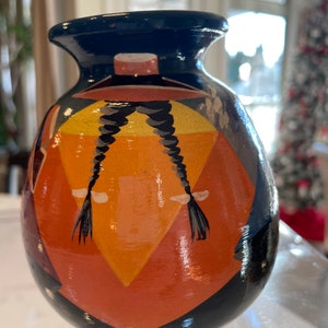 May include: A small, hand-painted ceramic vase with a geometric design in shades of orange, yellow, black, and blue. The vase features a stylized figure with braided hair.