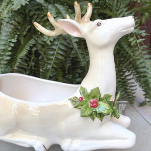 May include: A glossy cream-colored ceramic deer planter in a reclining position. The deer features gold-tipped antlers and a sculpted green holly leaf and red berry accent on its chest. The hollow body serves as a container for plants.
