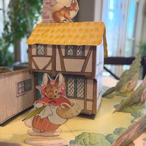 May include: A whimsical paper diorama featuring a storybook scene. A two-story house with a thatched roof is topped with a squirrel. A rabbit figure with a basket and umbrella stands nearby. The scene is set on a yellow landscape.