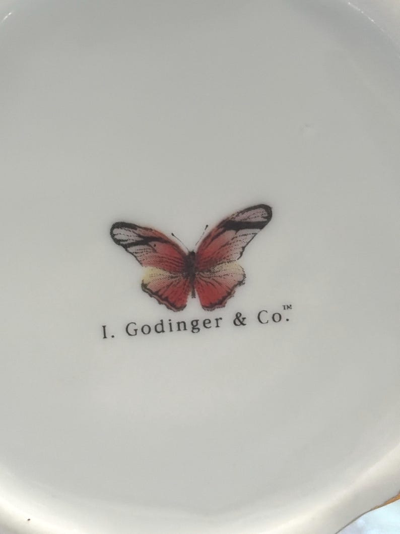 May include: White ceramic dish with a butterfly design in shades of red, pink, and black. The text "I. Godinger & Co." is printed below the butterfly. The dish has a slightly scalloped edge.