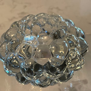 May include: A clear glass decorative bowl with a bubbly, textured surface. The bowl is round and appears to be made of individual, rounded glass pieces, creating a unique, organic shape. The bowl is sitting on a clear surface.