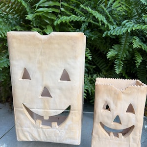 May include: Two off-white ceramic luminaries shaped like paper bags with cut-out jack-o'-lantern faces. The taller luminary is on the left and the shorter one is on the right. Both feature triangular eyes, a triangular nose, and a wide, toothy grin.