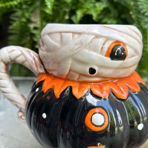 May include: A ceramic Halloween-themed mug. The top of the mug is designed to look like a mummy's head, with beige bandages and orange eyes. The bottom is a black pumpkin with orange accents and white polka dots. The handle is beige.