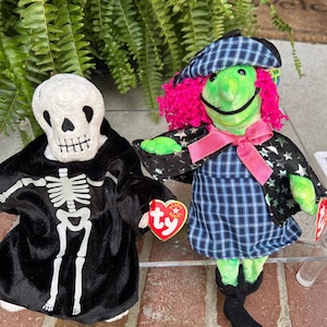 May include: Two Ty Beanie Babies: a skeleton in a black robe and a green witch with pink hair, a plaid hat, and a pink bow. The skeleton has a white skull face and a black robe with a white skeleton design. The witch wears a blue plaid dress.