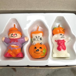 K&ouml;nnte beinhalten: Ein Set aus drei Halloween-Kerzen. Eine Kerze ist eine Figur in Lila und Orange, eine andere eine Katze auf einem K&uuml;rbis und die dritte ein Geist mit einer roten Schleife. Die Kerzen befinden sich in einem wei&szlig;en Tablett.