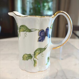 May include: White porcelain creamer with a gold rim and handle. The creamer is decorated with green leaves and a blue butterfly. The handle and rim are trimmed with gold. The creamer has a scalloped edge.