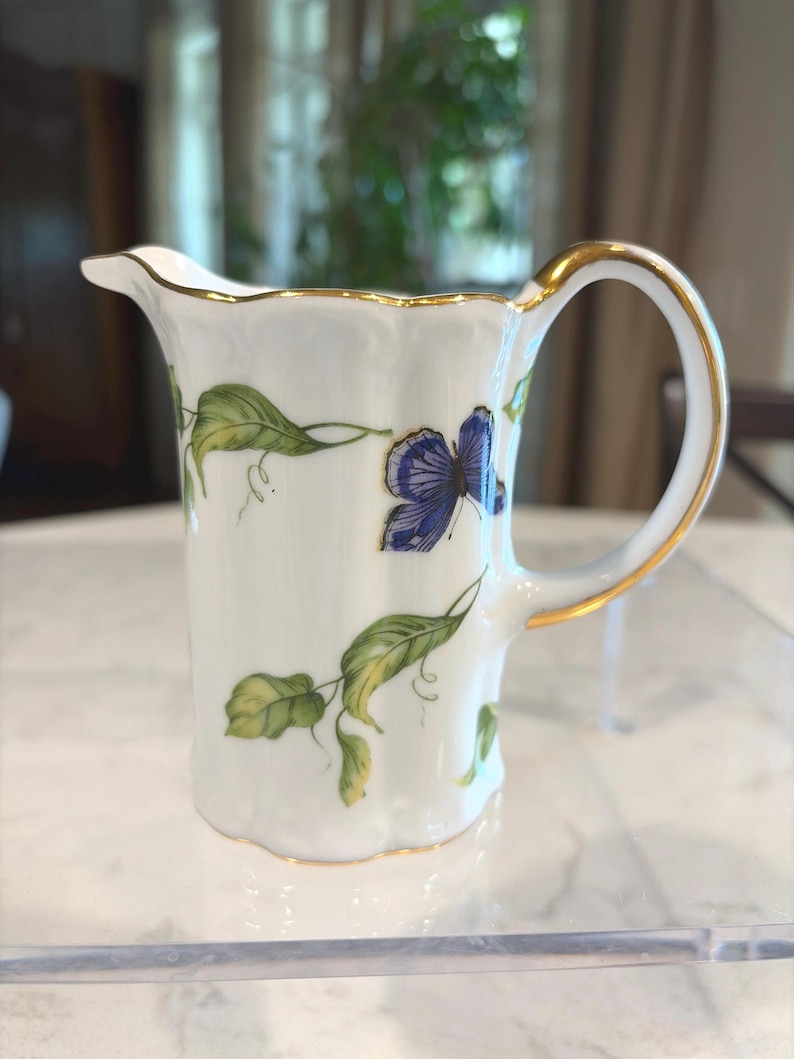 May include: White porcelain creamer with a gold rim and handle. The creamer is decorated with green leaves and a blue butterfly. The handle and rim are trimmed with gold. The creamer has a scalloped edge.