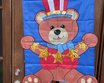 May include: A blue fabric flag featuring a brown teddy bear wearing a red, white and blue hat and holding gold stars. The bear is wearing a red shirt and a blue bow tie.
