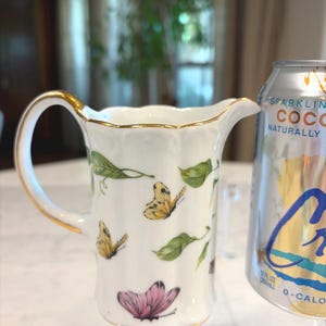 May include: White porcelain creamer with a gold handle and trim, decorated with butterflies and green leaves. The creamer has a spout for easy pouring. A can of sparkling coconut water is next to the creamer.