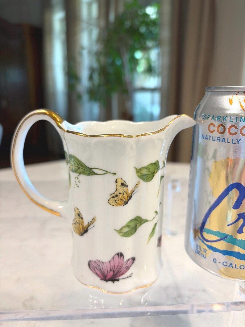 May include: White porcelain creamer with a gold handle and trim, decorated with butterflies and green leaves. The creamer has a spout for easy pouring. A can of sparkling coconut water is next to the creamer.