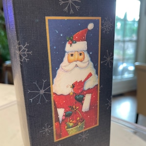 May include: A blue box with a white snowflake pattern. The front of the box features a festive illustration of Santa Claus holding a basket of fruit and a cardinal bird. The illustration is set against a blue background with white snowflakes.