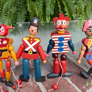 May include: Four vintage wooden jumping jack puppets featuring a jester, a soldier, a clown, and a pirate. Each figure has articulated limbs connected by strings with a red and white cord at the bottom to move the arms and legs when pulled.