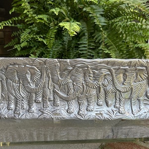 May include: A rectangular, silver-colored decorative tray featuring a detailed relief of a herd of elephants in a jungle setting. The elephants are depicted with intricate textures and are surrounded by foliage.
