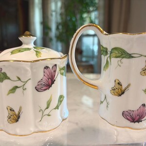 May include: A white porcelain sugar bowl with a lid and a creamer pitcher. Both are decorated with pink and yellow butterflies, green leaves, and gold trim. The sugar bowl has a gold finial on the lid.