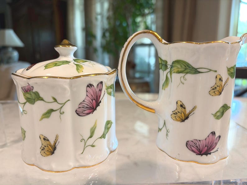 May include: A white porcelain sugar bowl with a lid and a creamer pitcher. Both are decorated with pink and yellow butterflies, green leaves, and gold trim. The sugar bowl has a gold finial on the lid.