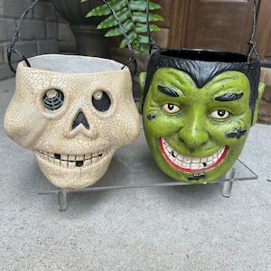 May include: Two Halloween treat buckets, one shaped like a skull with a spiderweb design and the other shaped like a green monster with a toothy grin.