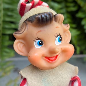 May include: A vintage Christmas elf ornament with a smiling face, blue eyes, and a red and white striped hat. The elf has a tan face and a red and white striped outfit. The ornament is designed for holiday decoration.