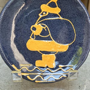 May include: A blue ceramic plate with a yellow Santa Claus design. The Santa Claus is wearing a hat and has a bag on his back. The plate has a wavy design around the edge.