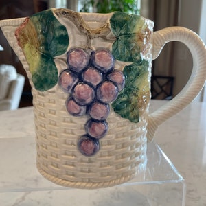 May include: Cream-colored ceramic pitcher with a woven basket design. The pitcher features a cluster of purple grapes and green leaves. The handle is white and twisted. A decorative piece for serving beverages.