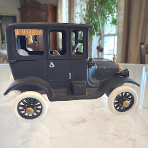 May include: A black antique car model with white-walled tires and gold accents. The vintage vehicle features a detailed interior and a classic design, perfect for collectors or as a decorative piece. The car is displayed on a glass surface.